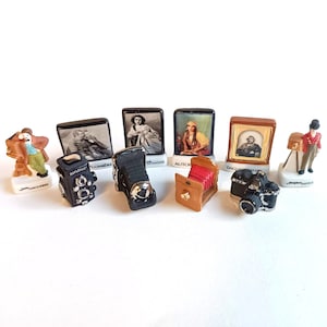 以下が含まれることがあります： ヴィンテージカメラと写真家の肖像画を描いたミニチュアセラミックフィギュアのコレクション。フィギュアには、さまざまなカメラモデル、カメラを持った写真家、有名な写真家の肖像画が含まれています。フィギュアは白い表面にあります。