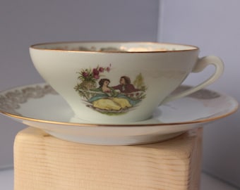 May include: A white porcelain teacup and saucer set with a gold rim. The cup features a painted scene of a couple in a garden setting.
