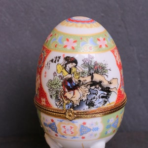 Puede incluir: Una caja de porcelana en forma de huevo con un borde dorado y tres patas doradas. La caja está decorada con un diseño floral colorido y una escena de una mujer con ropa tradicional.