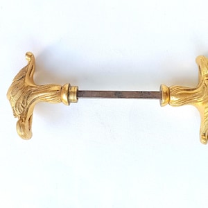 Peut inclure: Une poignée de porte dorée avec des motifs de têtes de lion sculptées sur chaque extrémité. La poignée est reliée par une tige en métal foncé. La quincaillerie est probablement vintage ou antique.