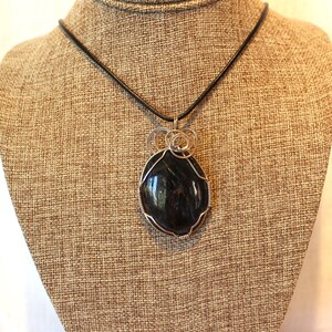May include: A black gemstone pendant necklace with silver wire wrapping. The pendant is oval shaped and hangs from a black leather cord.