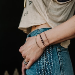 May include: A person wearing a light brown t-shirt with the word "Circle" on it, and blue jeans with stitching. They are wearing a set of four bracelets on their wrist, two are gold, one is black, and one is copper.