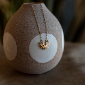 May include: A gold-colored necklace with a sunburst pendant is displayed on a textured, beige vase with white circular accents. The necklace chain is delicate, and the sunburst pendant has a detailed, spiky design.