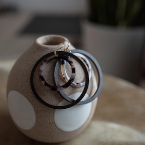 Könnte beinhalten: Ein Paar schwarze Holz-Creolen mit einem kleineren Schildpatt-Creolenring im Inneren. Die Ohrringe liegen auf einer beige-weißen, gepunkteten Vase.