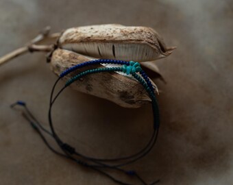 Può includere: Due braccialetti di perline con perline blu scuro e verde acqua. Un braccialetto ha piccole perline blu scuro e l'altro ha piccole perline verde acqua. I braccialetti sono su una superficie marrone strutturata.