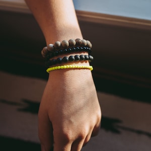 Puede incluir: Una muñeca con cuatro pulseras apiladas una encima de la otra. La pulsera superior está hecha de cuentas de madera marrón. La segunda pulsera está hecha de cuentas negras. La tercera pulsera está hecha de cuentas negras. La pulsera inferior está hecha de cuentas de color amarillo neón.