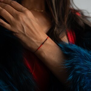 May include: A delicate bracelet featuring a string of small, red and black beads. The bracelet is worn on a wrist, with a red string and a clasp. The background includes a blue fur coat and a red top.