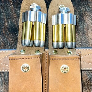 May include: Two brown leather belt loops with metal snaps, each holding a silver and brass bullet holder with five bullets.