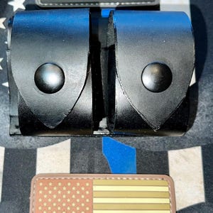 May include: A black leather double pouch with two snap button closures, positioned between two rectangular patches. The top patch is a black and grey American flag with a thin blue line, and the bottom patch is a tan and brown American flag.