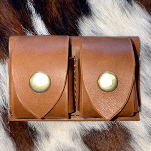 May include: A brown leather double pouch with two metal snaps. The pouch has a flap on each side and is designed to hold small items.