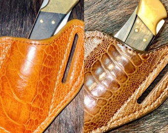 May include: A folding pocket knife with a brass bolster and dark wood handle, partially inserted into a tan leather sheath featuring a textured, ostrich-like grain pattern and a vertical belt loop slot for secure carrying.