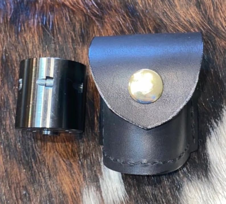 Może przedstawiać: Srebrny metalowy cylinder w czarnym sk&oacute;rzanym etui. Etui ma zapięcie na zatrzask i srebrny guzik.