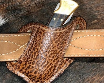 May include: A brown leather knife sheath with a stitched belt loop. A folding knife with a brass handle and wood inlay is partially inserted into the sheath.