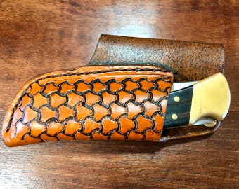May include: A brown leather knife sheath with a patterned design. The sheath has a flap that folds over the knife. The knife has a brass handle and a black wooden handle.