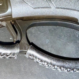 May include: Close-up of a black firearm trigger and trigger guard. The trigger guard has a stitched leather grip. The trigger is silver. The firearm is set against a dark background.