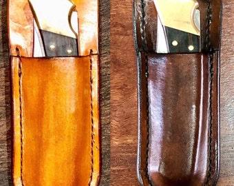 May include: Two folding pocket knives with dark wood handles and brass bolsters, each housed in a custom-fitted leather sheath. The left sheath is light tan leather, while the right sheath is dark brown leather, both featuring brass snap closures.
