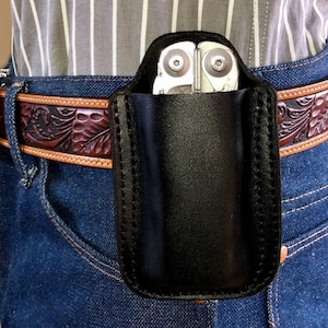 May include: A black leather belt holster for a multi-tool. The holster is attached to a brown leather belt with an intricate floral design. The multi-tool is visible inside the holster.