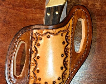 May include: A brown leather knife sheath with intricate tooling and a decorative border. The sheath has two belt loops and is designed to hold a folding knife.