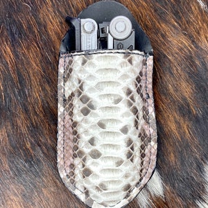 May include: A white and brown snakeskin leather pouch with a black leather trim. A multi-tool is partially visible inside the pouch.