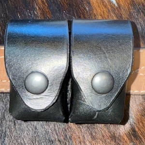 May include: Two black leather pouches with silver snap closures are attached to a light brown leather belt. The pouches are rectangular with rounded tops. The background is a mix of brown and black animal hide.