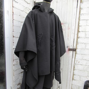 May include: A black hooded cloak and trousers are displayed on a mannequin. The cloak has decorative patches and a flowing design. The outfit is made of a dark material, possibly wool. The mannequin is positioned outdoors against a white brick wall and a white door.
