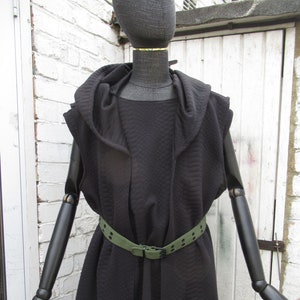 May include: A black hooded vest with a quilted pattern, worn with a green belt. The vest is sleeveless and has a draped design. The mannequin is black with articulated arms and hands.