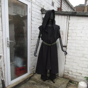 May include: A black hooded cloak and matching pants are displayed on a mannequin. The cloak has a textured pattern and is cinched at the waist with a green belt. The mannequin stands in front of a white brick wall and a white door.