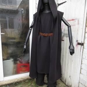 May include: A black hooded cloak and matching pants displayed on a black mannequin. The cloak has a quilted texture and is cinched at the waist with a brown belt. The mannequin has articulated arms and is positioned outdoors.