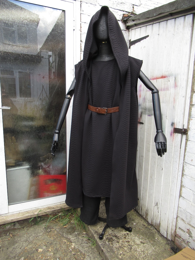 May include: A black hooded cloak and matching pants displayed on a black mannequin. The cloak has a quilted texture and is cinched at the waist with a brown belt. The mannequin has articulated arms and is positioned outdoors.