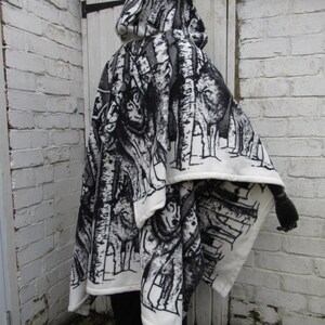 May include: A white hooded fleece blanket with a black and white print of trees and wolves. The blanket is folded in half and draped over a person's shoulders.