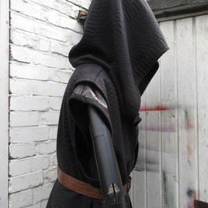 May include: Black hooded cloak with quilted texture, leather accents on the shoulders, and a brown leather belt. The cloak is displayed on a black mannequin arm against a white brick wall and a white door.