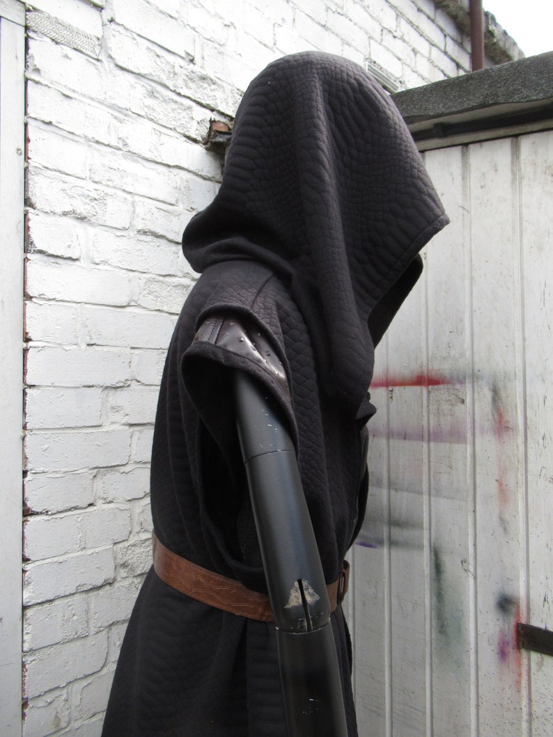 May include: Black hooded cloak with quilted texture, leather accents on the shoulders, and a brown leather belt. The cloak is displayed on a black mannequin arm against a white brick wall and a white door.