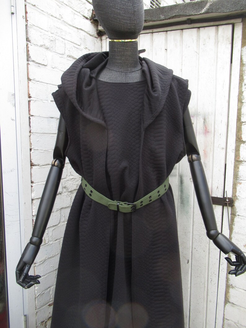 May include: A black hooded vest with a quilted pattern, worn with a green belt. The vest is sleeveless and has a draped design. The mannequin is black with articulated arms and hands.