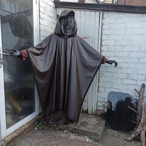 May include: A dark brown hooded cloak with a subtle textured pattern, worn over a brown long-sleeved garment. The cloak features a wide, flowing silhouette with black trim along the edges, paired with black gloves and a grey-haired mannequin head.