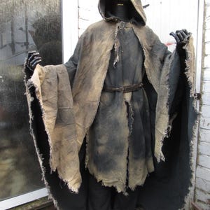 May include: A dark costume with a hooded cloak and tattered edges. The cloak is a mix of black and tan, with a belt at the waist. The costume includes black gloves and trousers, creating a spooky, medieval look. The date 05/03/2026 is visible.