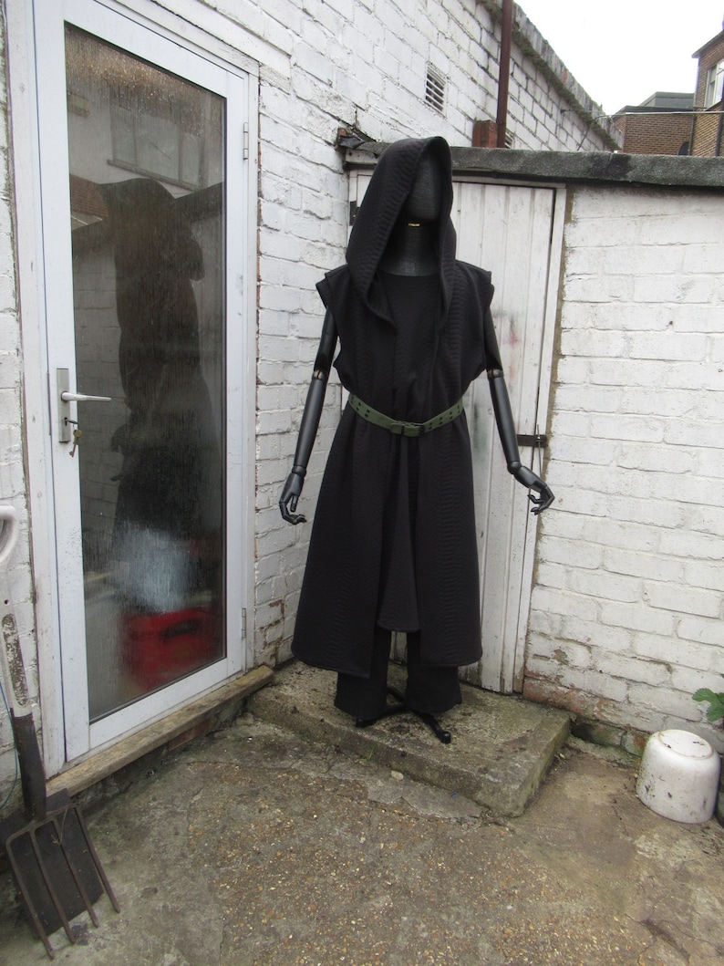 May include: A black hooded cloak and matching pants are displayed on a mannequin. The cloak has a textured pattern and is cinched at the waist with a green belt. The mannequin stands in front of a white brick wall and a white door.