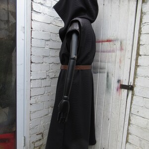 May include: A black hooded cloak with a quilted texture, featuring short sleeves and a brown belt. The cloak is displayed on a black mannequin against a white brick wall and a white door.