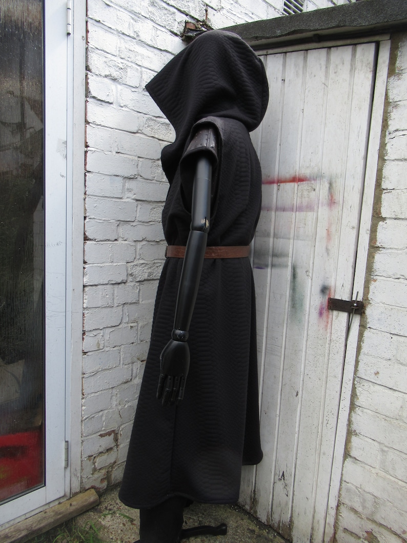 May include: A black hooded cloak with a quilted texture, featuring short sleeves and a brown belt. The cloak is displayed on a black mannequin against a white brick wall and a white door.