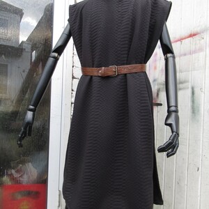 May include: Black quilted tunic dress with a brown leather belt and a silver buckle. The dress has a unique textured pattern and a sleeveless design. The mannequin has black articulated arms and is displayed outdoors.