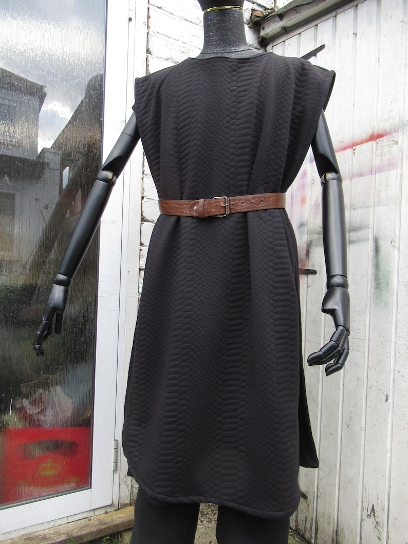 May include: Black quilted tunic dress with a brown leather belt and a silver buckle. The dress has a unique textured pattern and a sleeveless design. The mannequin has black articulated arms and is displayed outdoors.