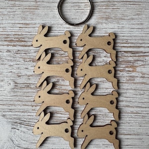May include: A collection of eight wooden bunny cutouts with a silver metal ring. The bunnies are light brown, with a simple design and a small hole. The ring is at the top, and the bunnies are arranged on a light wood-grain surface.