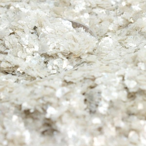 May include: Close-up of a pile of iridescent, off-white mica flakes. The flakes have a shimmering, textured appearance, reflecting light. The mica appears to be in a loose, granular form, suitable for crafts or decorative purposes.