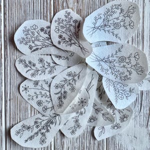 May include: A collection of irregular, off-white fabric patches featuring black line art illustrations of various wildflower bouquets and botanical stems. Each patch displays unique floral arrangements with delicate petals, leaves, and thin stems.