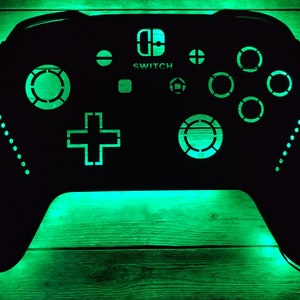 May include: Black controller-shaped wall art with green LED lighting. The controller design includes the word "SWITCH" and the Nintendo Switch logo. The controller is mounted on a wooden background.