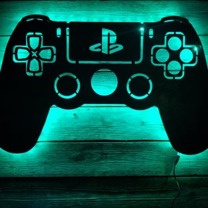 May include: A black metal PlayStation controller silhouette with a teal green light behind it. The controller is mounted on a wooden wall.