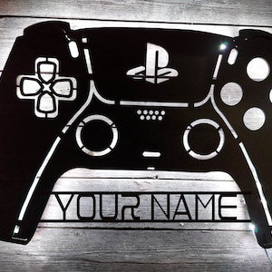 May include: Black metal cutout of a PlayStation controller with the PlayStation logo and the text "YOUR NAME" underneath. The controller is lit from behind, creating a glowing effect.