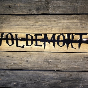 May include: Black metal sign with the word "VOLDEMORT" in a stylized font, featuring a wand design. The sign is mounted on a rustic wooden background, with a warm light illuminating the text.
