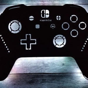 May include: Black wall art shaped like a video game controller, featuring the word "SWITCH" and controller buttons. The controller design has a series of small white lights along the edges. The background is a wooden surface.