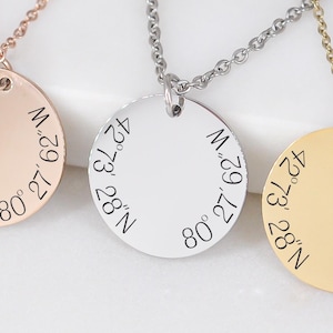 Puede incluir: Tres collares con colgantes redondos en oro rosa, plata y oro. Cada colgante tiene una cadena y coordenadas grabadas: 42°73 28°N 80°27 62°W. Los collares se muestran sobre una superficie blanca con cuentas de madera en el fondo.