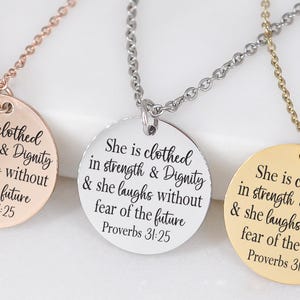 Könnte beinhalten: Drei Halsketten mit runden Anhängern. Die Anhänger sind roségold, silber und goldfarben. Jeder Anhänger trägt die Inschrift "She is clothed in strength & Dignity & she laughs without fear of the future Proverbs 31:25."